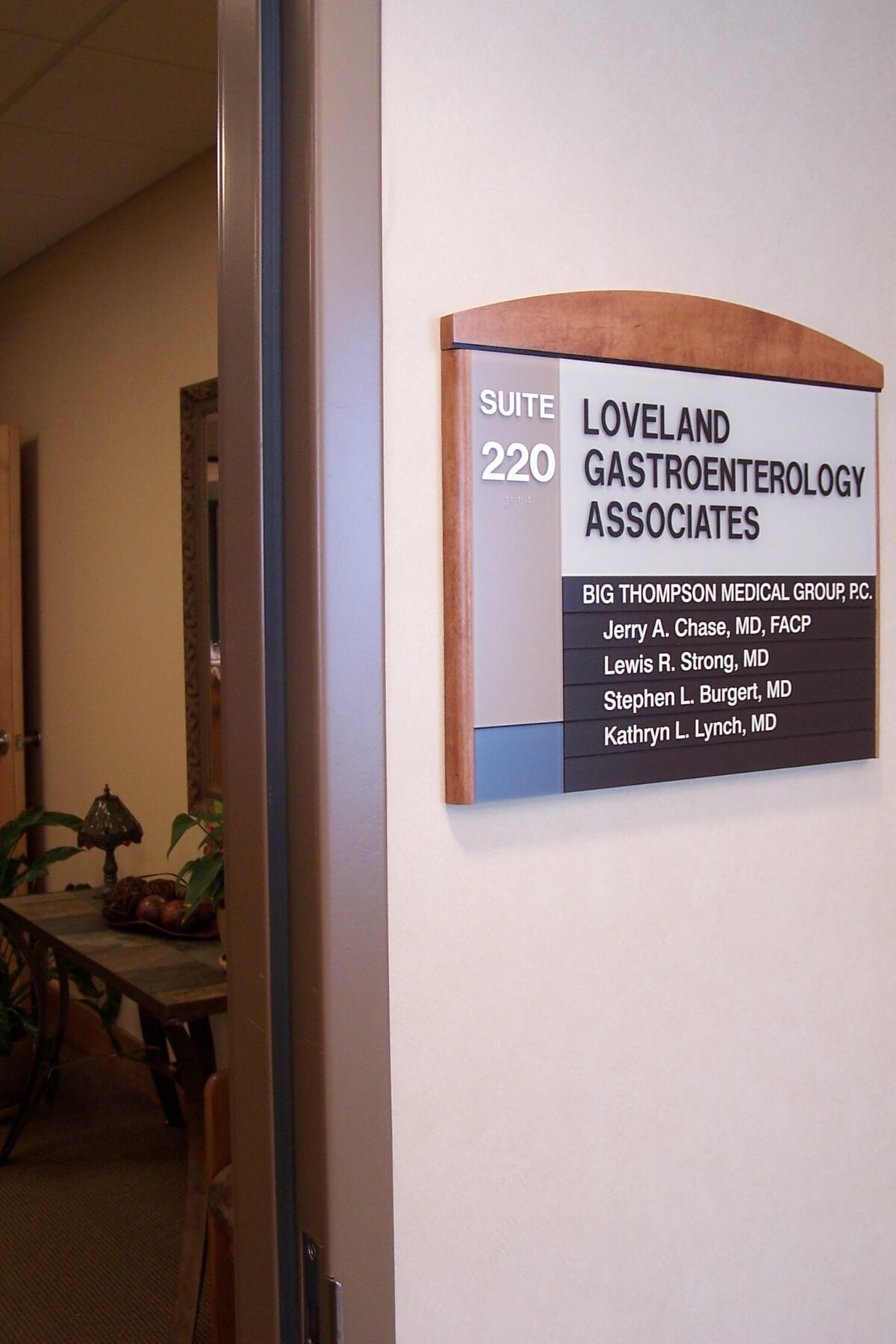 A sign on a wall reading "Suite 220, Loveland Gastroenterology Associates" with names of several doctors underneath. The sign is next to an open door leading into an office with plants and furniture visible inside.