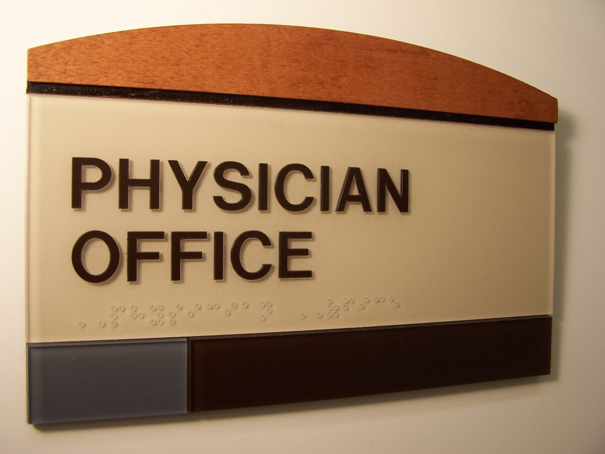 A wall-mounted sign with a wooden arch at the top reads "PHYSICIAN OFFICE" in large, bold letters. The sign consists of beige, brown, and gray panels with Braille text beneath the main lettering.