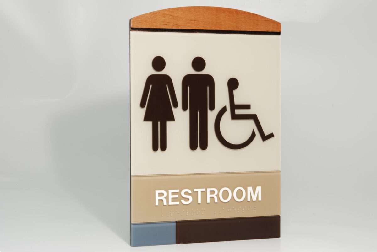 Sign indicating a restroom for multiple users. The sign has pictograms of a woman, a man, and a person in a wheelchair, above the text "RESTROOM". The top of the sign is curved and colored in brown and beige tones.