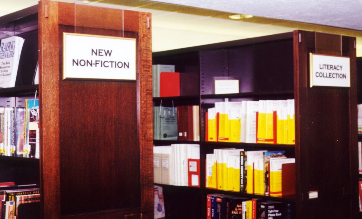 Two wooden bookshelves in a library. The left shelf, labeled "New Non-Fiction," is filled with various books. The right shelf, labeled "Literacy Collection," contains books with yellow spines and other materials.