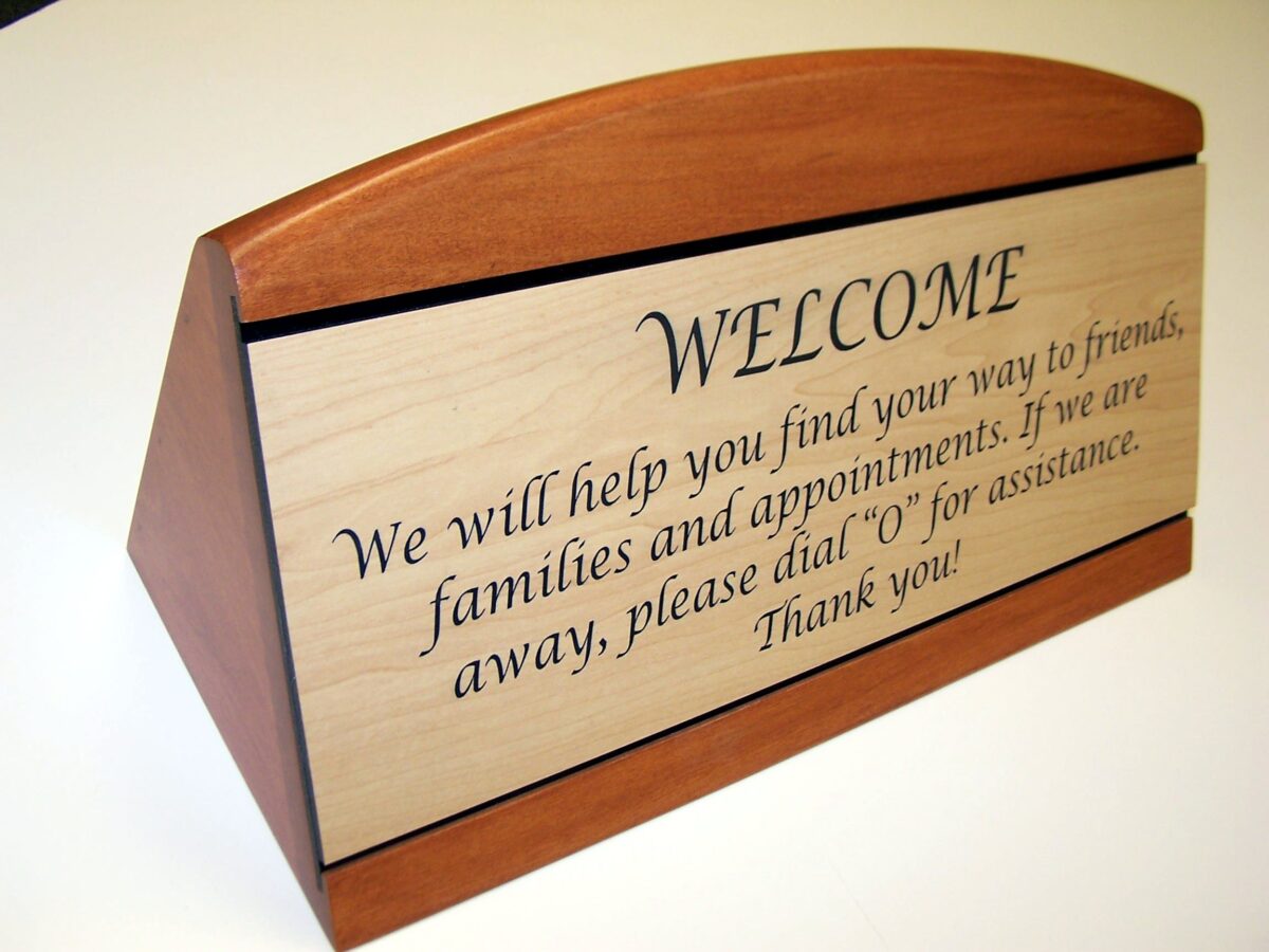 A wooden desk sign with a light-colored front panel engraved with a welcome message in elegant script. The message reads, "WELCOME. We will help you find your way to friends, families and appointments. If we are away, please dial '0' for assistance. Thank you!.
