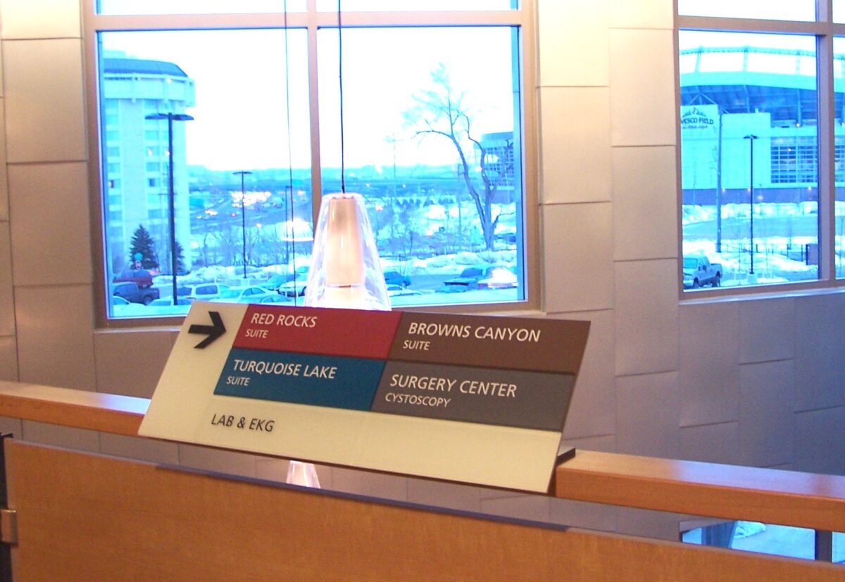 A directional sign inside a building points to various locations: to the right for Browns Canyon and Surgery Center/Orthopedic, and to the left for Red Rocks and Turquoise Lake. Large windows in the background reveal a snowy, urban landscape.