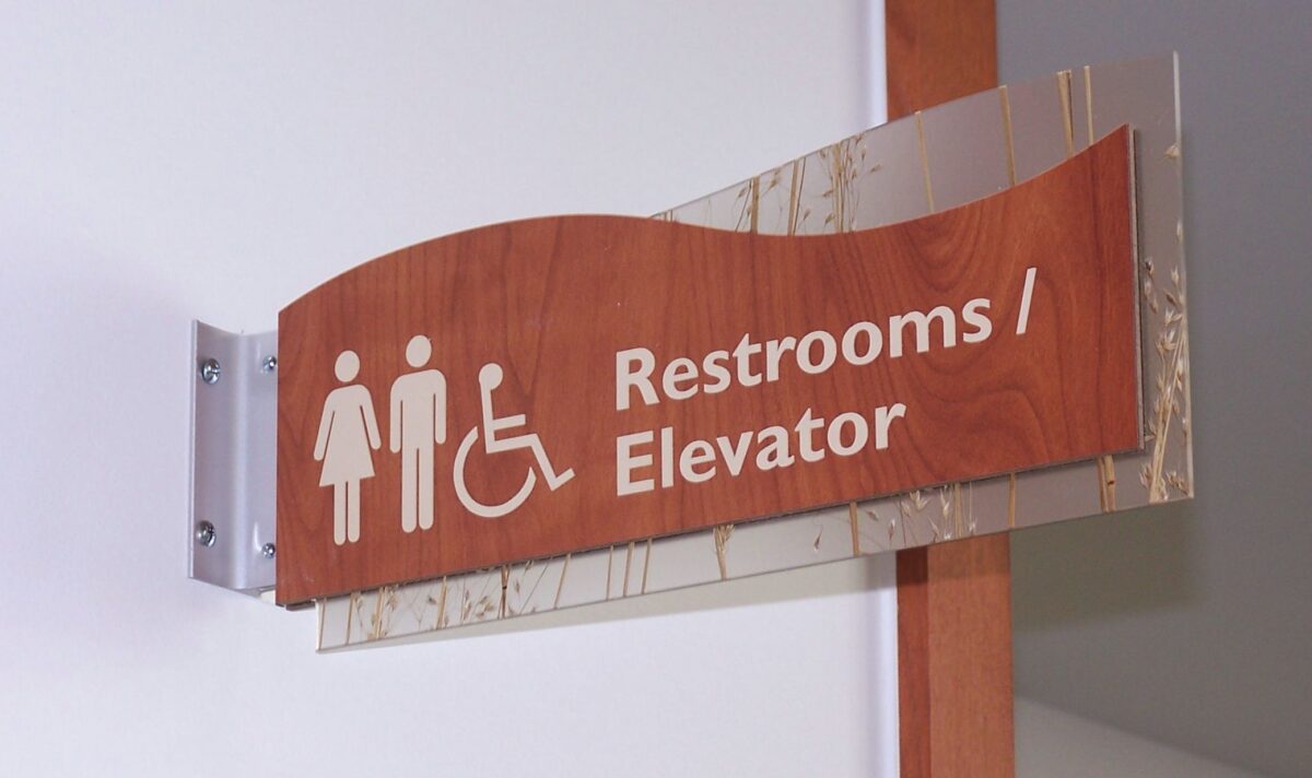 A wooden sign with white icons and text indicates the locations of restrooms and an elevator. The sign features symbols for men, women, and individuals with disabilities. The sign is mounted on a wall in a hallway.