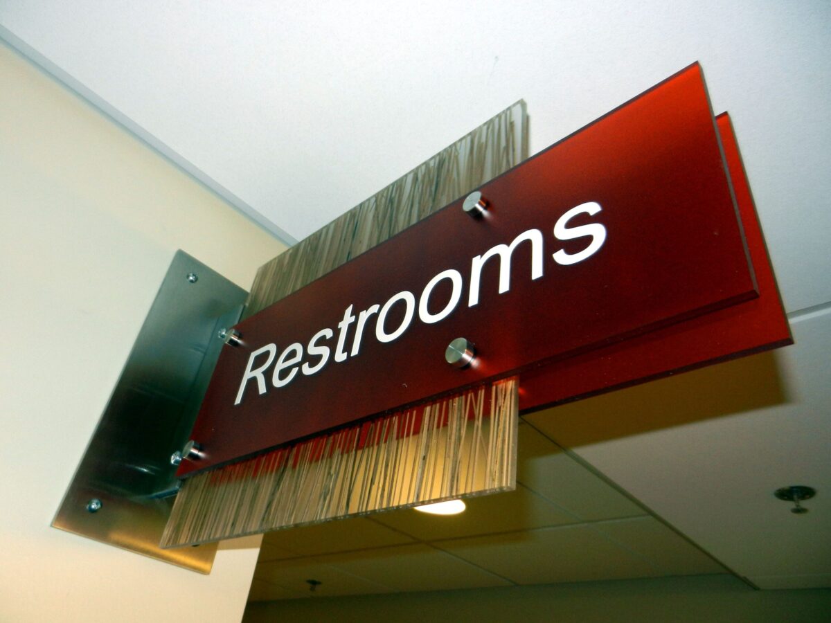 A directional sign for restrooms is mounted on a wall. The sign is red with white text that reads "Restrooms." The sign also includes a decorative element with vertical lines on the left side. It is lit from above by a ceiling light.