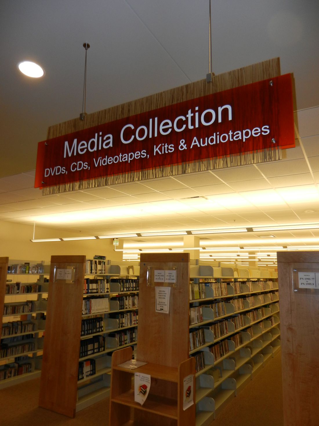 A library's media collection section with a sign hanging from the ceiling that reads "Media Collection: DVDs, CDs, Videotapes, Kits & Audiotapes." Below the sign are tall shelves filled with various media items. The area is well-lit with overhead lighting.