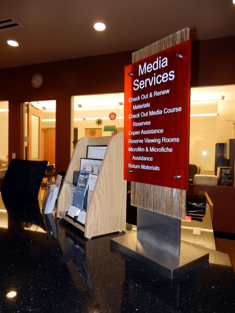 A library media services desk with a red sign listing available services including course reserves, media check-out and renewals, and group assistance. Brochures and information are displayed in holders on the counter. The background shows additional office space.