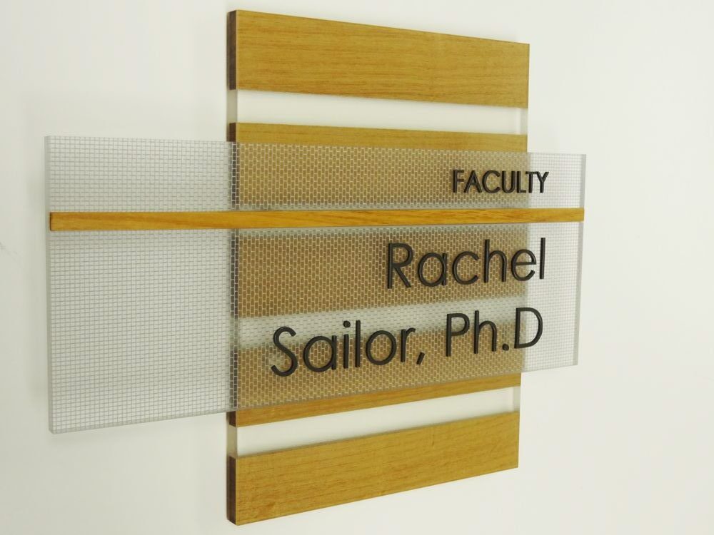 A wall-mounted sign with wood and frosted glass panels reads "FACULTY" at the top and "Rachel Sailor, Ph.D" below it in black text. The sign is placed on a white wall.