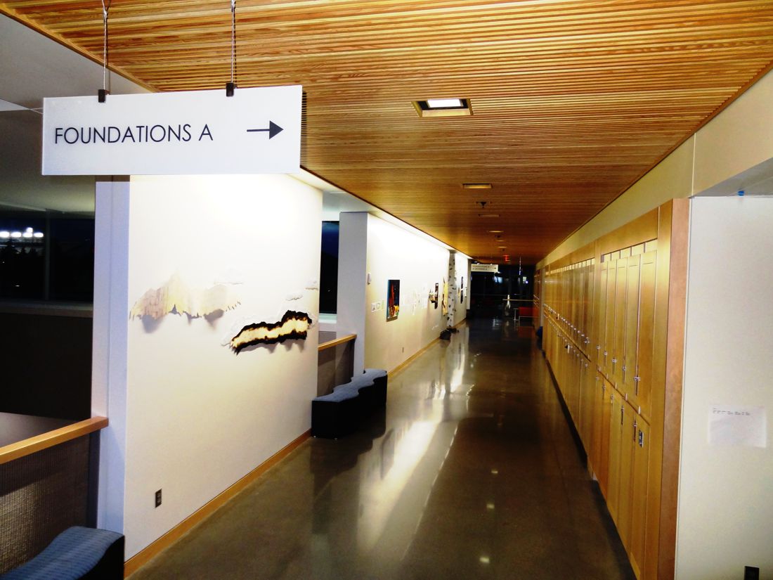 A brightly lit hallway with wooden ceiling and lockers on the right. The floor is polished concrete. Art pieces hang on the left wall, and a sign overhead reads "Foundations A" with an arrow pointing to the right.