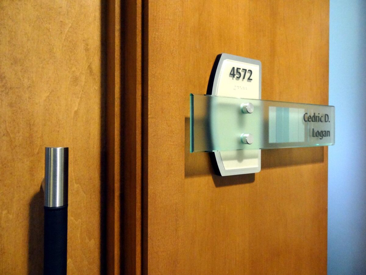A close-up image shows a wooden door with a contemporary metal handle on the left side. Attached to the door is a modern nameplate with the number "4572" and the name "Godric D. Legrande" written on it. The nameplate is accentuated with a clear glass panel.