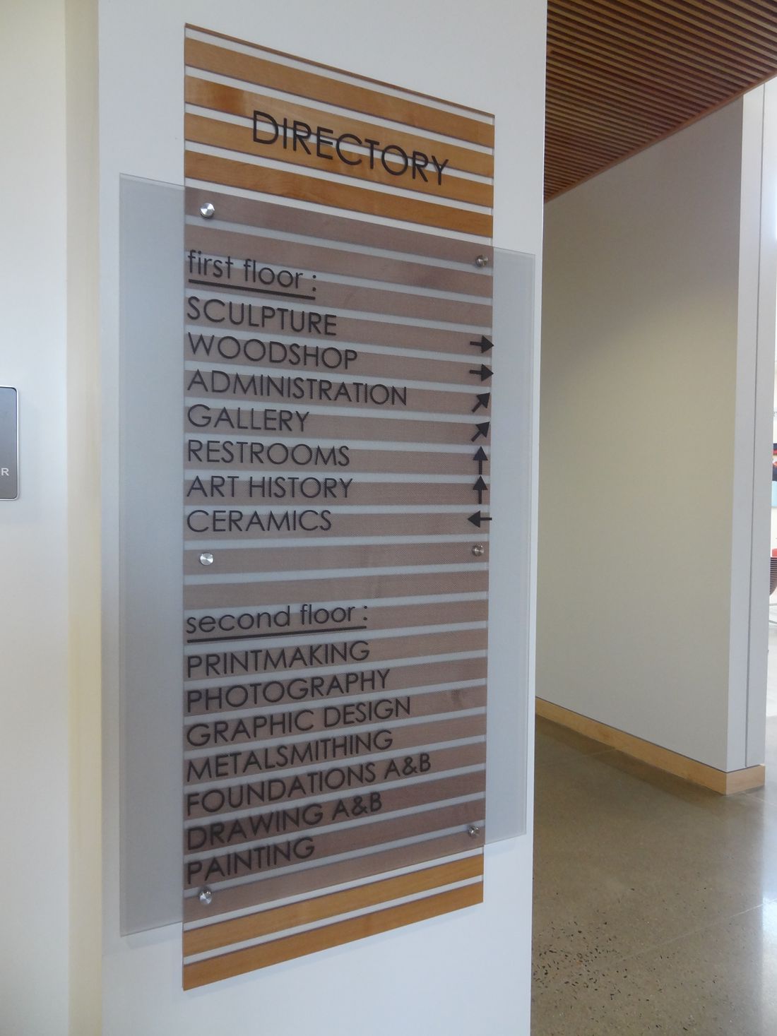 A directory sign in a building lists various locations: Sculpture, Woodshop, Administration, Gallery, Restrooms, Art History, and Ceramics on the first floor; Printmaking, Photography, Graphic Design, Metalsmithing, Foundations A&B, Drawing A&B, and Painting on the second floor.