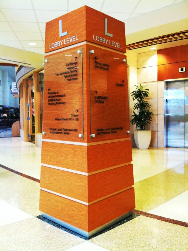 A wooden directory pillar stands in a lobby area, featuring a large "L" at the top for "Lobby Level." The directory lists various departments and offices in black text on clear panels. The surroundings include a potted plant, elevator doors, and a glossy tiled floor.