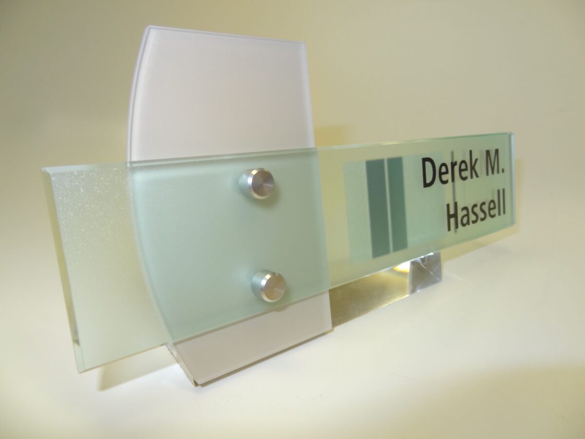 A translucent, modern nameplate with frosted glass and metallic accents reads "Derek M. Hassell." The nameplate features a contemporary design with a combination of clear and frosted glass segments, secured by two metal bolts.