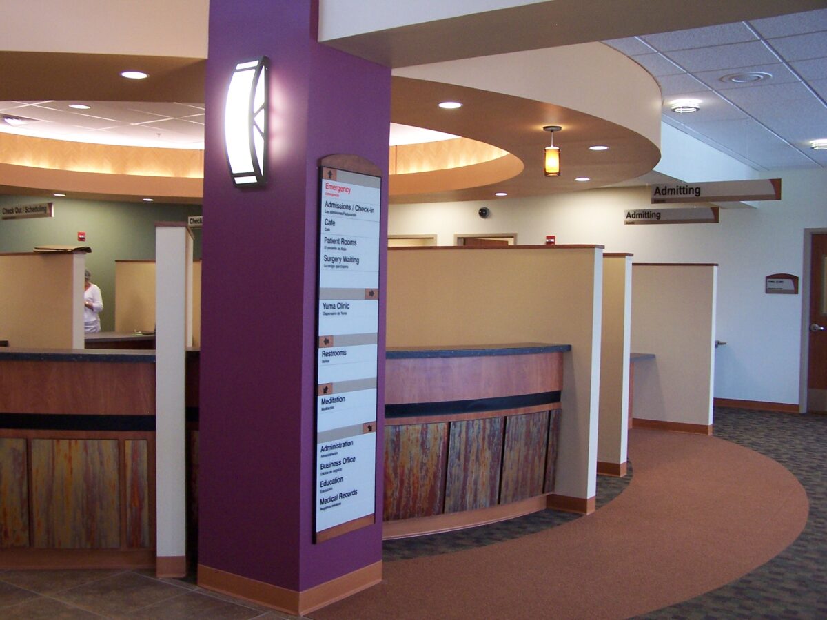 A modern medical reception area featuring a curved wooden desk with dividers, a purple column with a light fixture and a sign listing departments. Neutral-toned walls and carpeting provide a calming atmosphere. Signs point to various sections of the facility.