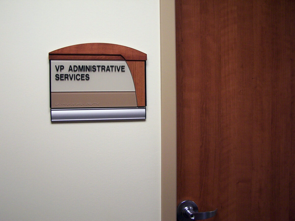 A wall-mounted sign next to a wooden door reads "VP Administrative Services." The sign has a brown and beige color scheme with a silver bottom section. The door is partially visible with a metal handle.