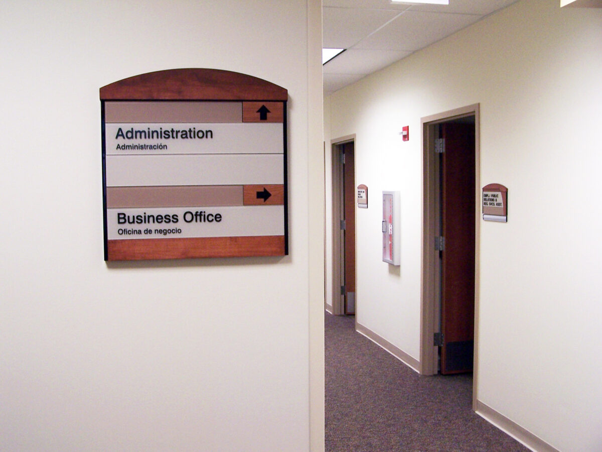 A hallway in an office building with beige walls and carpet. A sign on the left wall has two sections, one pointing up for “Administration” and one pointing right for “Business Office.” The hallway has several doors and is lit by ceiling lights.