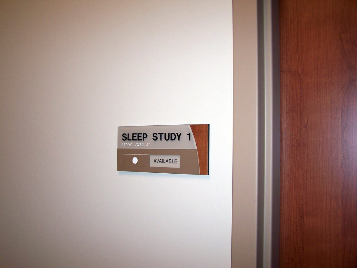 A sign on a light-colored wall reads "Sleep Study 1." Below the text, there is a small rectangular light with the word "AVAILABLE" illuminated in green. To the right of the sign is a wooden door.