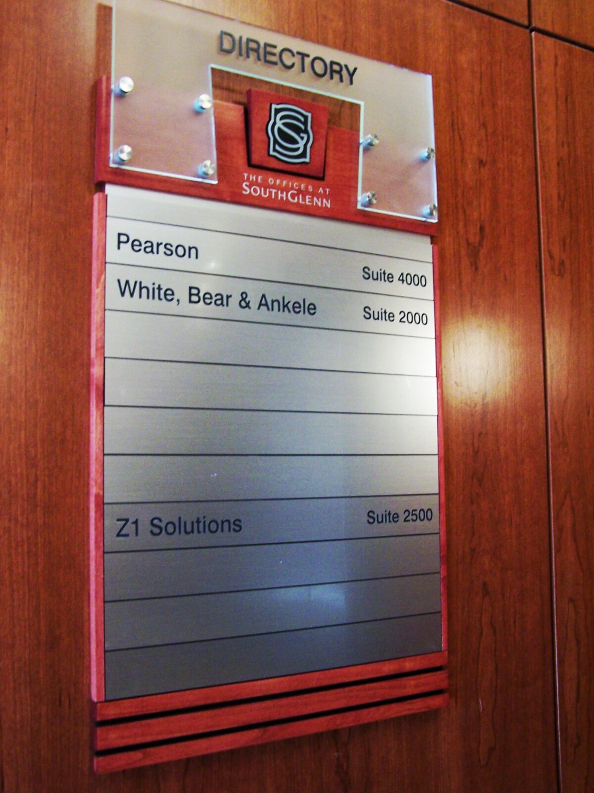 A wall-mounted directory sign with a clear header displaying the "University of South Glenn" logo. Below, it lists three entities: "Pearson" in Suite 4000, "White, Bear & Ankele" in Suite 2000, and "Z1 Solutions" in Suite 2500. The background is wooden.