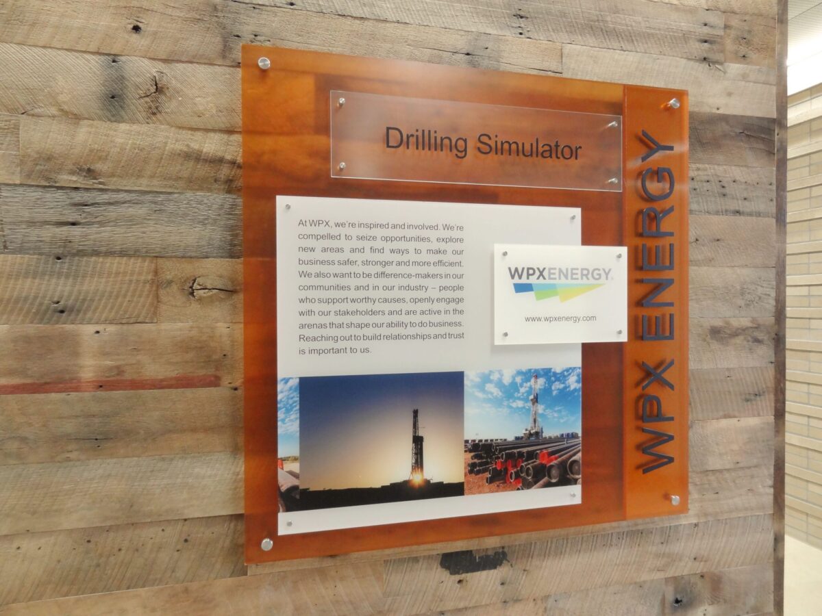 A sign mounted on a wooden wall reads "Drilling Simulator" at the top. Below, a description with the WPX ENERGY logo is displayed, along with two images: one of a drilling rig at sunset and another of a drilling operation. The WPX ENERGY logo is also on the side.