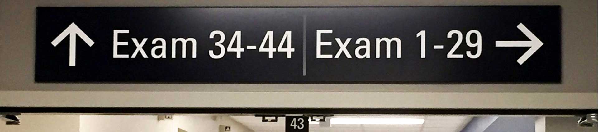 A long, horizontal sign in a hospital corridor indicating directions to various exam rooms, with arrow symbols pointing right for "Exam 1-29" and left for "Exam 34-44," set against a neutral background to ensure clarity and ease of navigation