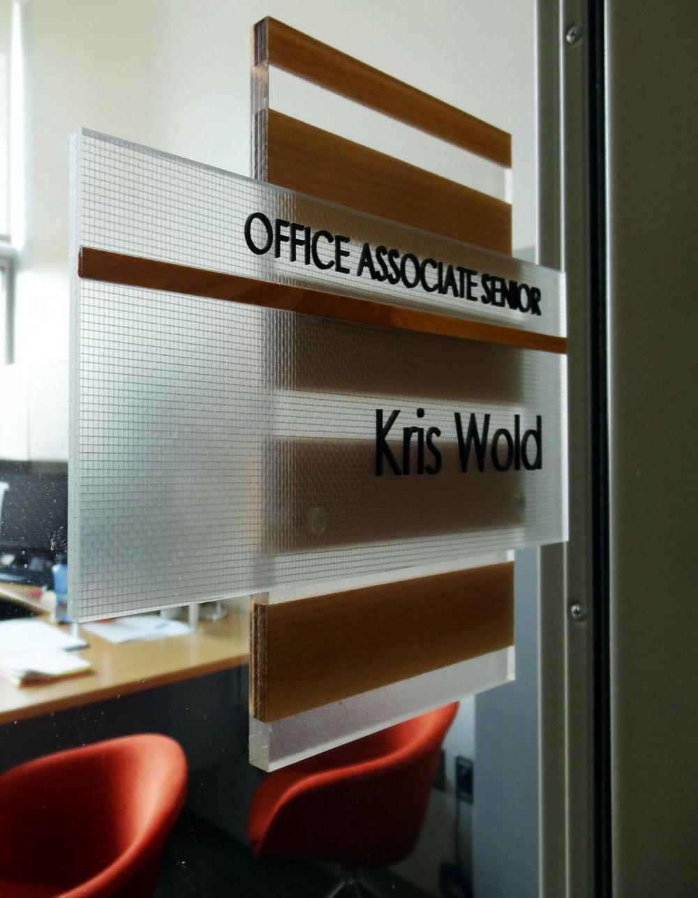 Office door sign for 'Kris Wold, Office Associate Senior' displayed in a clear and wooden frame on a glass door, with an office interior visible in the background.