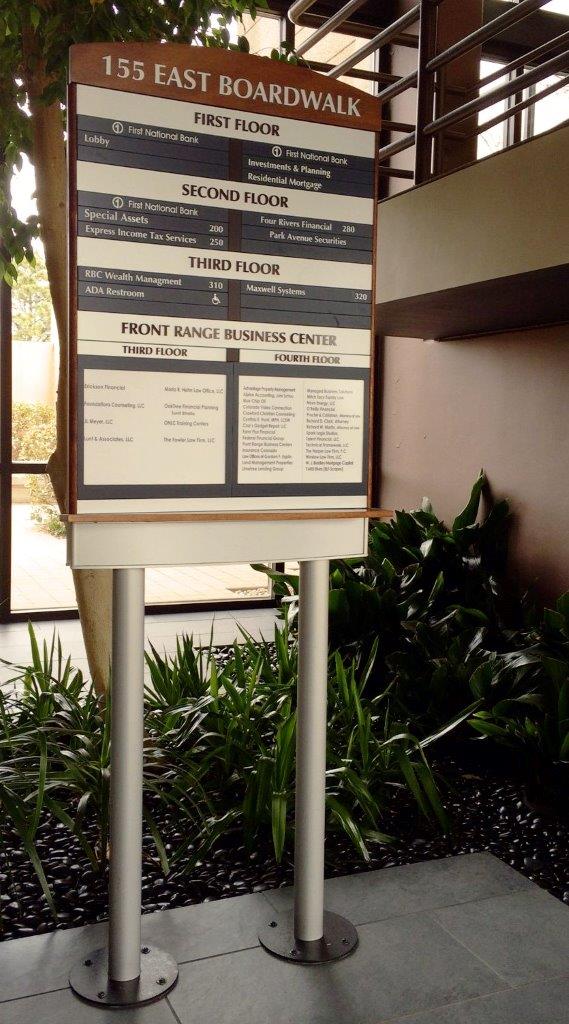 Directory sign at 155 East Boardwalk showing floor listings for various businesses, standing in an indoor setting with surrounding potted plants and a background of windows and railings.