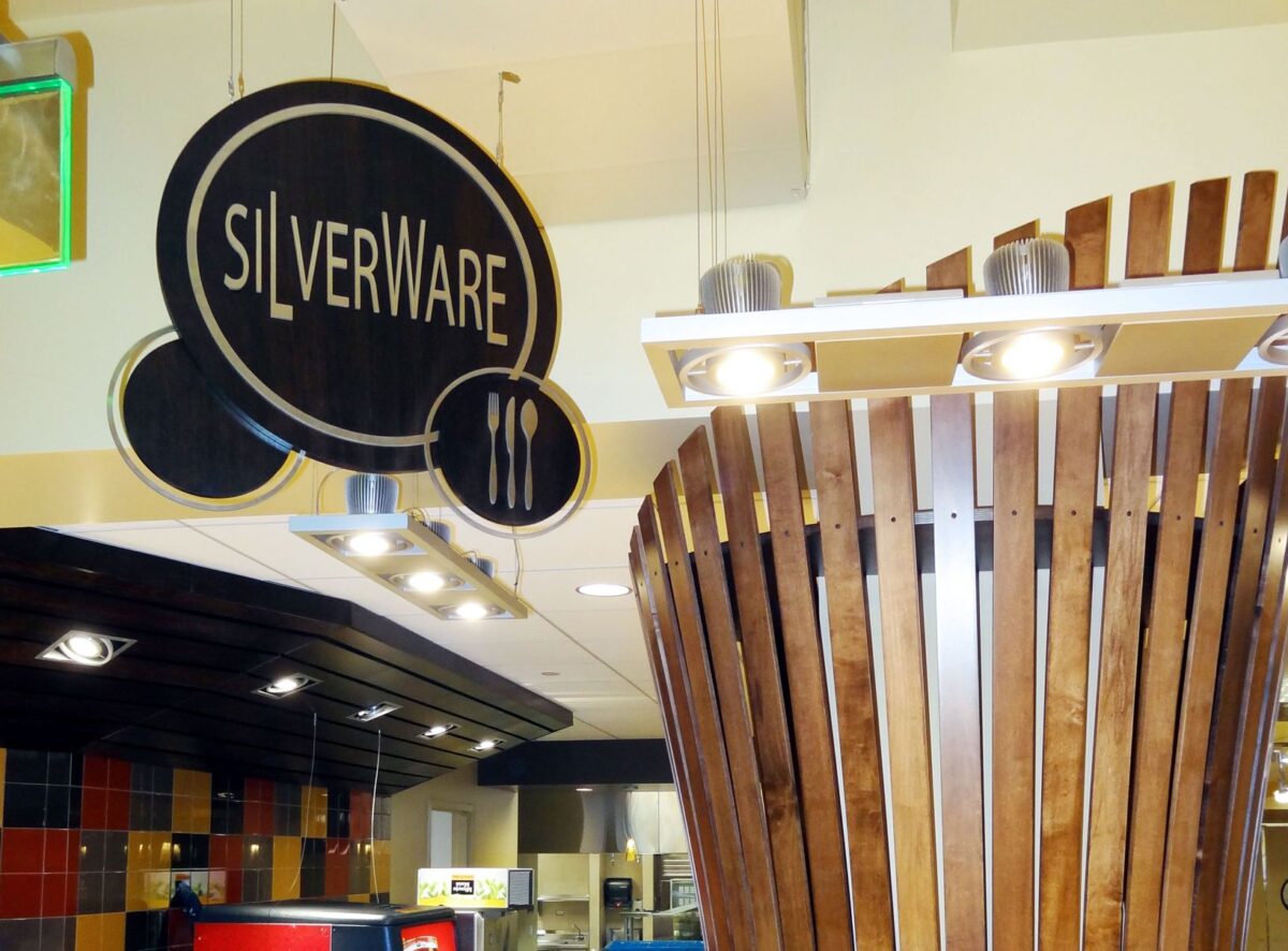 A dining area sign reading "SILVERWARE" hangs from the ceiling, featuring a fork, knife, and spoon icon. In the background are colorful tiles and a ceiling fixture designed with vertical wooden slats.
