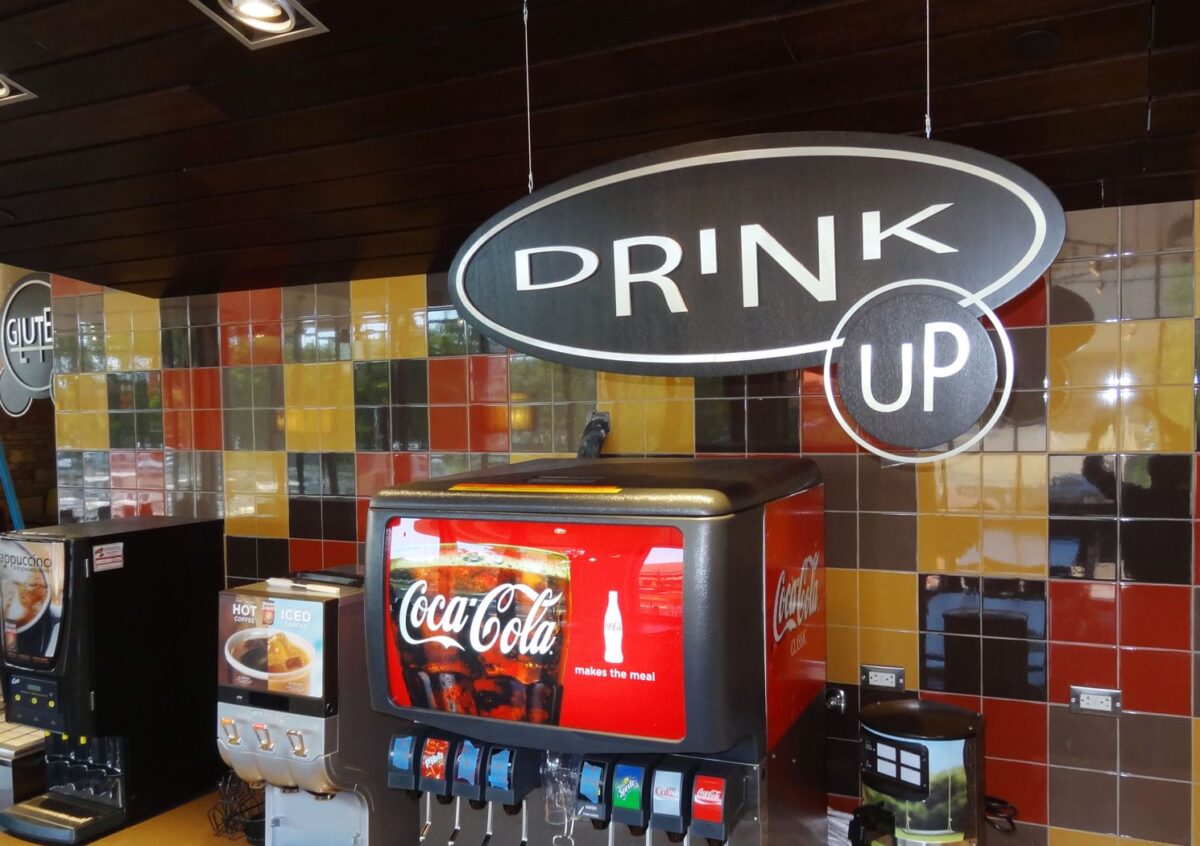 Self-service beverage station in a restaurant with a Coca-Cola dispenser. The station has a variety of drink options under a large sign reading "DRINK UP." The background features a colorful, tiled wall.