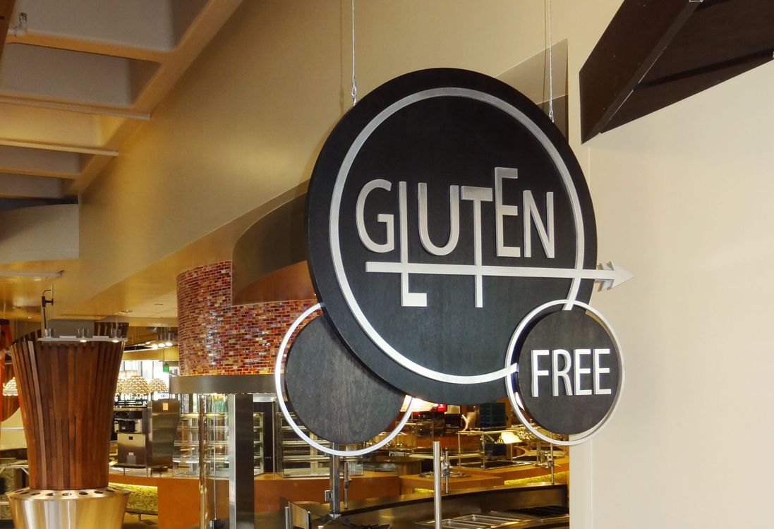 A black circular sign hangs in a restaurant, displaying the words "GLUTEN FREE" in white, modern font. The background of the restaurant shows blurred images of the counters and food displays.