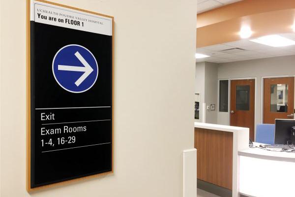 Directional sign inside a hospital indicating exam room locations and exit routes, mounted on a wall with an arrow pointing right.