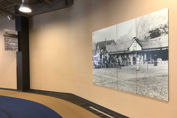 A large black-and-white historical photograph hangs on a light beige wall in a spacious room with high ceilings. The floor has a curving path design with a combination of blue and tan tiles. The photo depicts a group of people standing outside old buildings.
