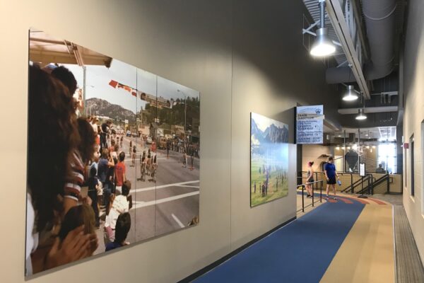 A spacious indoor hallway features large photos on the walls. On the left is an image of people observing an outdoor event. The hallway has overhead lights and a blue and beige walkway.