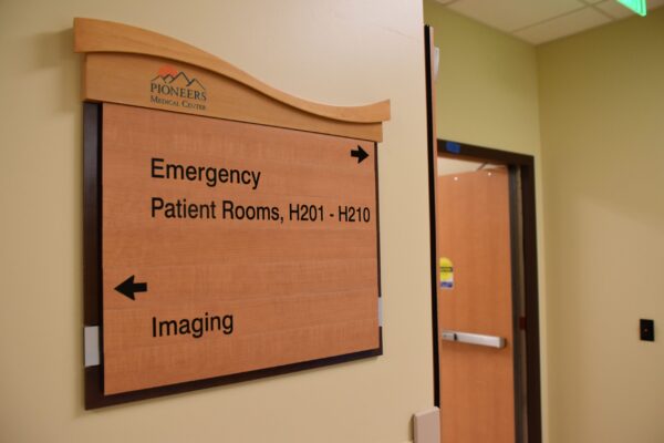 A directional sign inside a building points left for the Emergency and Patient Rooms H201-H210, and right for Imaging. The sign hangs on a beige wall next to an open wooden door. The ceiling lights and a portion of the corridor are visible.