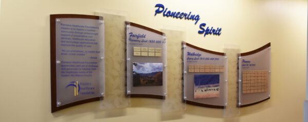 A wall display titled "Pioneering Spirit" features four panels with text, images, and charts. The first panel has text and a logo, the second and third include images and charts, and the fourth has a chart. The background wall is painted a light beige color.