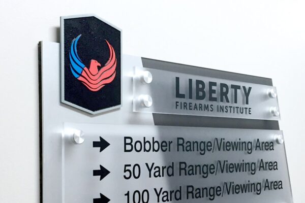 A wall-mounted sign with a black and red emblem at the top left, and "Liberty Firearms Institute" written next to it. Below, arrows point right to three different locations: "Bobber Range/Viewing Area," "50 Yard Range/Viewing Area," and "100 Yard Range/Viewing Area.