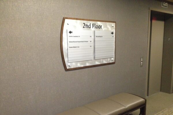 A hallway with a directory sign mounted on a grey wall indicating "2nd Floor." The sign lists various room numbers and details. Below the sign is a brown bench with a cushioned seat. To the right, there is an elevator door and a control panel.