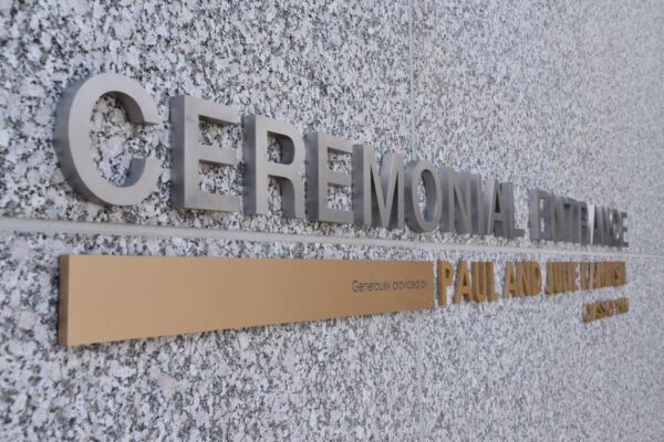 A wall with large stainless steel letters spelling "CEREMONIAL ENTRANCE" and a smaller, horizontal brass plaque underneath. The wall surface is speckled gray. The angle of the photo is slightly tilted, providing a side view of the text.