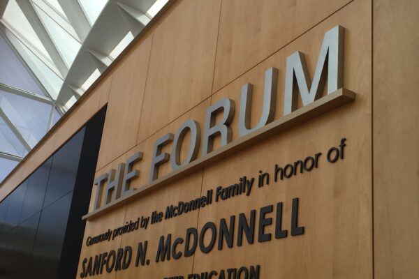 A wooden wall with mounted metal letters reading: "THE FORUM." The wall also displays the text: "Generously provided by the McDonnell Family in honor of Sanford N. McDonnell, A World Leader in Character Education." Brightly lit ceiling visible in the background.