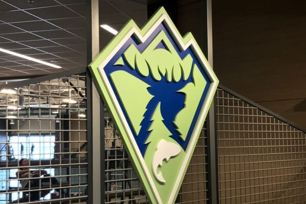 A logo features a stylized blue moose head with antlers above a leaping fish, against a green and blue mountain backdrop. The logo is mounted on a metal fence-like structure inside a building with industrial elements and visible ceiling grid.