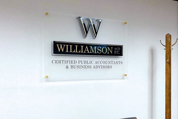 A sign on a wall reads "Williamson & Co PC, Certified Public Accountants & Business Advisors" below a metallic logo with the letter "W". Two gray chairs with wooden armrests are in front of the sign, and a wooden coat rack is on the right side.
