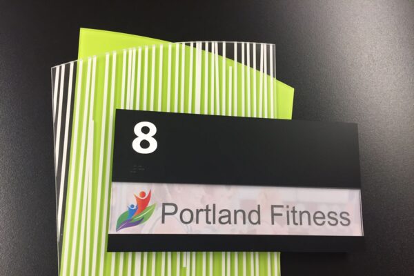 A black rectangular sign with the number "8" in white at the top. Below it, there is a colorful logo of three human figures and the text "Portland Fitness" on a white background. The sign is set against a background of vertically striped green and white paper.