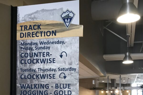A sign in a gym indicates track directions: counter-clockwise on Monday, Wednesday, Friday, and Sunday; clockwise on Tuesday, Thursday, and Saturday. Walking lanes are blue and jogging lanes are gold. Below the sign, the track is visible with fitness equipment nearby.