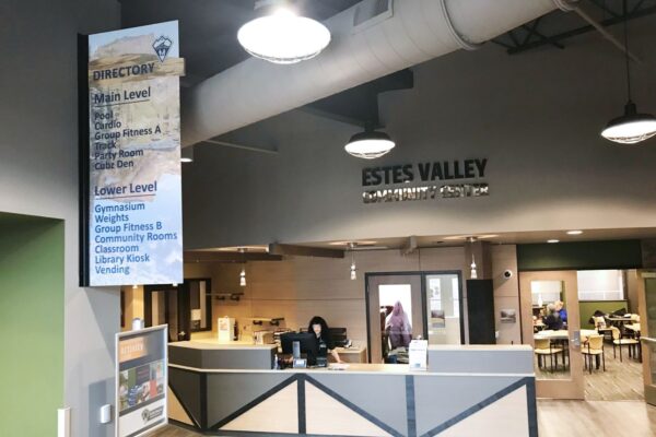 Lobby of Estes Valley Community Center featuring a reception desk with a staff member, a large directory sign listing amenities, and a prominent ceiling sign with the center's name. Background includes tables and chairs with people engaging in activities.