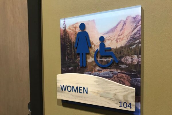 A restroom sign on a wall indicating a women's restroom, featuring symbols for women and accessibility for people with disabilities. The background includes a landscape image, and the sign is labeled "WOMEN" with the number "104" at the bottom.