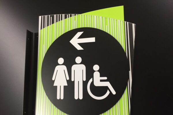 A restroom sign with icons indicating facilities for men, women, and people with disabilities. The sign is on a black circle with a left-pointing arrow and is set against a lime green background with white and black vertical stripes at the top.