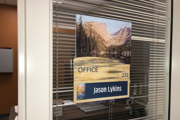 A glass door with blinds features a sign displaying a nature scene and the text "OFFICE 272 Jason Lykins." The office interior is visible through the window.