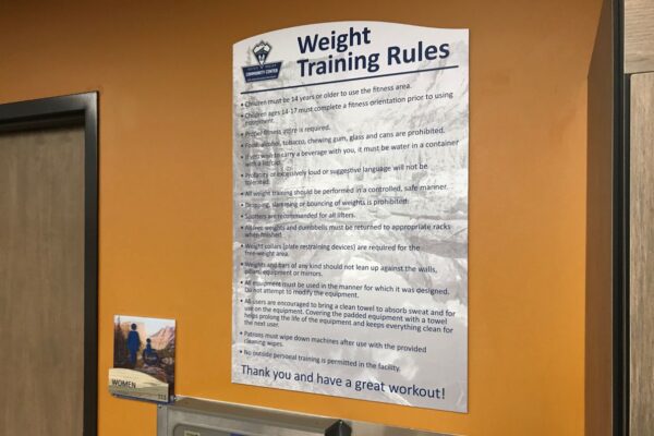Weight training rules sign posted on an orange wall near a doorway. The sign contains various guidelines for weight training, emphasizing safety, proper equipment use, and courtesy towards other gym users. Below the sign is a water fountain and a small notice.