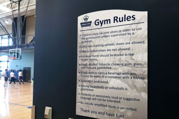 A gym rules sign is posted on a dark wall in an indoor basketball court. The rules specify not to bring gum, tobacco, food, or drinks, and highlight the requirement for appropriate attire and music. People are visible playing basketball in the background.