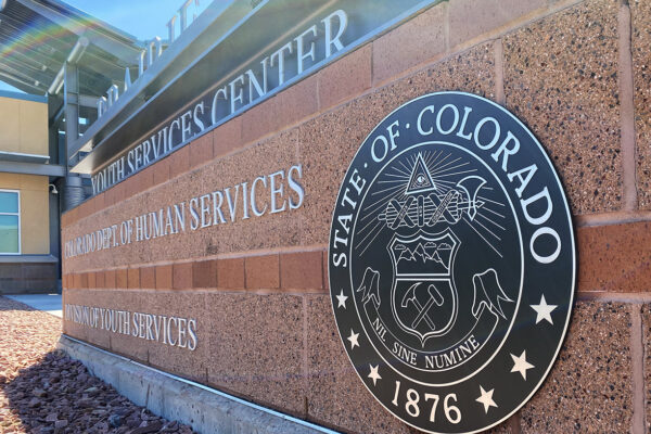 A brick wall with a large circular emblem that reads "State of Colorado 1876" and the text "Department of Human Services" and "Youth Services". The emblem has various symbols, including a pickaxe, a scroll, and a Roman fasces with an axe.