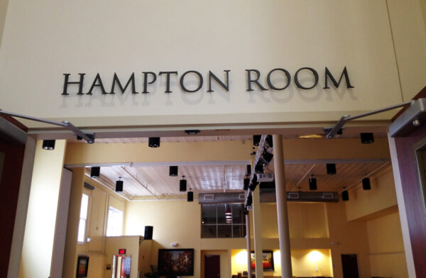 A wide-open doorway with the sign "Hampton Room" above it. Inside is a spacious, well-lit room with high ceilings, light-colored walls, and rows of black light fixtures hanging from the ceiling.