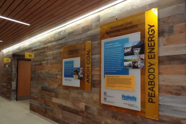 A hallway with wooden walls features two large informational posters. One poster is titled "Peabody Energy" and the other is titled "Arch Coal." Both posters contain text and images. There are doorways and directional signs also visible in the hallway.